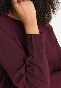 Blouse bordeaux à manches longues avec détails de boutons et fronces aux poignets, confectionnée dans un tissu doux et texturé.