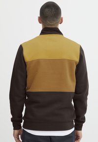 Fleece-Pullover mit hohem Kragen, ausgestattet mit gelb-braunen und dunkelbraunen Farbblocks, glatter Textur und gerippten Bündchen.