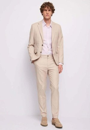 Jonge man met krullend haar, gekleed in een beige pak, wit overhemd en beige loafers, staand tegen een effen lichte achtergrond.