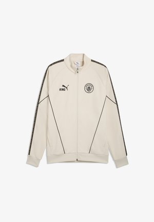 Beige Jacke mit durchgehendem Reißverschluss, schwarzer Paspelierung, Puma-Logo und "KING"-Schriftzug auf der rechten Brust, Manchester City Emblem auf der linken Brust, gemusterte Streifen an den Ärmeln.