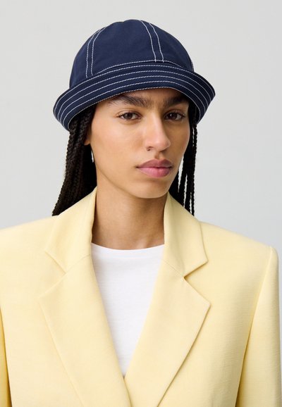 Cappello da pescatore blu con cuciture bianche, realizzato in tessuto morbido. Abbinato a un blazer giallo chiaro e una camicia bianca, presenta un look a strati.