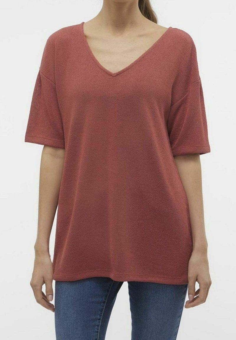 Femme portant un pull ample, texturé, à manches courtes et col en V couleur rouille, associé à un jean bleu sur un fond uni.