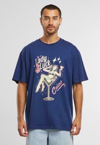 Sötétkék pamut póló rövid ujjakkal, melyen egy színes grafika látható, amely egy nőt ábrázol egy martini pohárban, valamint a "Lady Luck Casino" felirat.