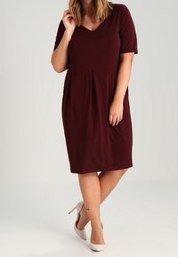 Abito bordeaux al ginocchio con scollatura a V, maniche corte e vita arricciata. Tessuto leggero con una texture liscia, abbinato a tacchi beige.