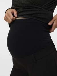 Grūtniece, kas paceļ melnu grūtniecības blūzi, ģērbusies melnās biksēs un mirdzošā melnā krekliņā uz neitrāla fona.