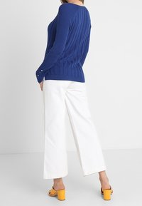 Niebieski prążkowany sweter z długimi rękawami i ozdobnymi guzikami, zestawiony z szerokimi białymi spodniami i żółtymi sandałami na obcasie typu blok.