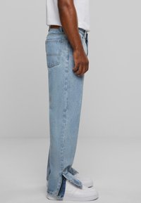 Jeans de mezclilla azul claro con corte recto, cremalleras laterales en los dobladillos y bolsillos traseros, combinados con zapatillas blancas y una camisa blanca.