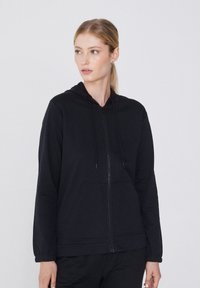 Haut à capuche zippé noir en tissu doux, doté d'une capuche, de poches avant et de manches longues avec des poignets élastiques. Design minimaliste.