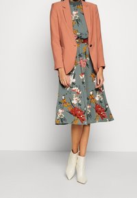 Blazer color blush sopra un abito floreale con motivi verdi, rossi e gialli. Indossato con stivali alla caviglia bianchi. Tessuto morbido e forma strutturata.