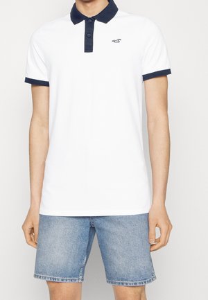 Mand iført en hvid polo-shirt med sort krave og ærmekant, kombineret med lyseblå denimshorts, står mod en hvid baggrund.
