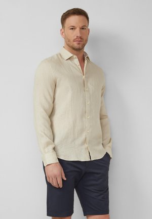 Mann trägt beige langärmliges Hemd mit Knöpfen und marineblaue Shorts, steht vor einfachem hellgrauem Hintergrund.