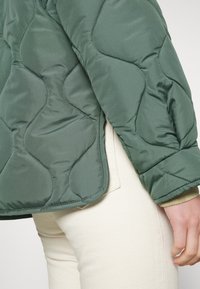 Giacca trapuntata verde con una texture esterna liscia, orlo arrotondato e polsini elastici. Indossata sopra pantaloni color crema.