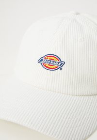 Boné de corduroy branco com um logótipo bordado da Dickies em azul, amarelo e vermelho. O boné tem uma forma estruturada com textura estriada.