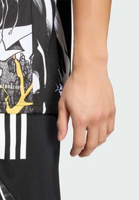 Zbliżenie na rękę przy czarno-białej sportowej koszulce z żółtymi akcentami oraz czarnych szortach z białymi paskami.