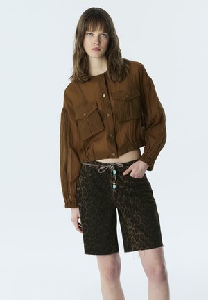 Młoda kobieta o prostych brązowych włosach, ubraną w brązową teksturyzowaną kurtkę zapinaną na guziki oraz ciemne, w panterkę, krótkie spodenki sięgające kolan.
