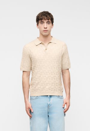 Νεαρός άνδρας φορώντας μπεζ πικέ κοντομάνικο πόλο και ανοιχτόχρωμο μπλε τζιν, στέκεται μπροστά σε ένα απλό ανοιχτό φόντο.