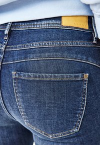 Dunkelblaue Denim-Jeans mit strukturiertem Finish, verziert mit kontrastierenden orangefarbenen Nähte und einem gelben Lederpatch am hinteren Bund.