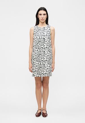 Γυναίκα με ίσια σκούρα μαλλιά που στέκεται, φοράει αμάνικο μαύρο-άσπρο φόρεμα με floral σχέδια και σκούρα κόκκινα παπούτσια Mary Jane σε λευκό φόντο.