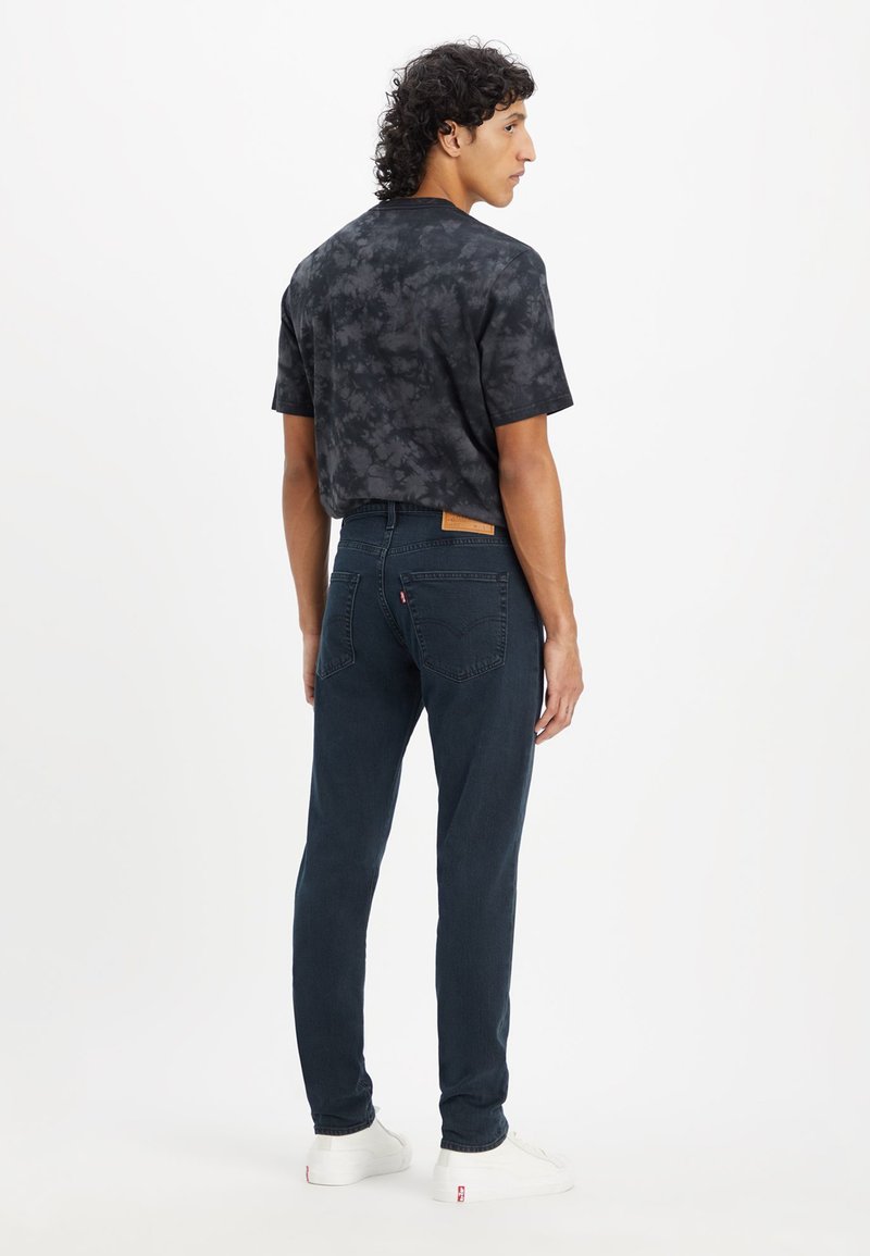 Chemise noire tie-dye à manches courtes associée à un jean bleu foncé et des baskets blanches. Le jean présente une étiquette en cuir à l'arrière.