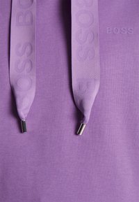 Sudadera morada con el logo "BOSS" en relieve y cordones a juego. Los cordones tienen terminales de metal plateado; la tela parece suave y sin costuras.