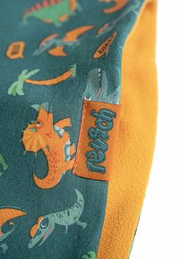 Farverigt dinosaurermønsteret stof i teal og orange, der har en blød tekstur med livlige illustrationer og et syet orange label.