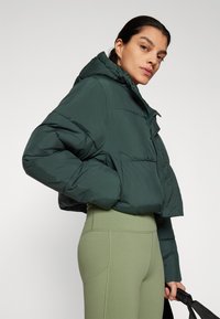 Veste courte en doudoune verte avec capuche, dotée d'une texture lisse, d'une fermeture éclair et de poches latérales, portée avec des leggings olive taille haute.