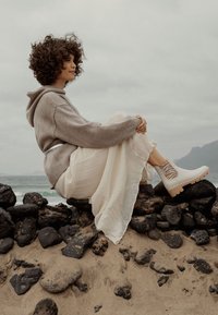 Sudadera oversize gris claro, falda blanca fluida y botas tipo botín de crema con un diseño texturizado. Sentada sobre piedras oscuras junto a la playa.