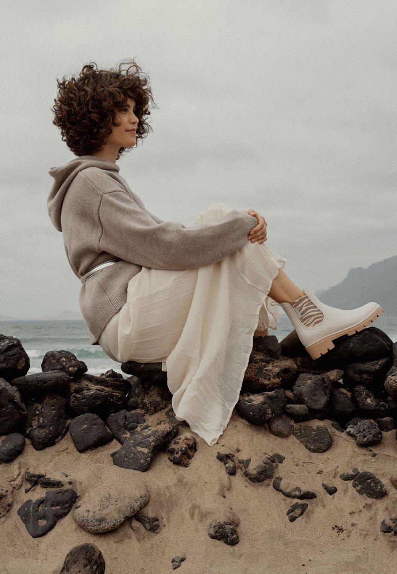 Sudadera oversize gris claro, falda blanca fluida y botas tipo botín de crema con un diseño texturizado. Sentada sobre piedras oscuras junto a la playa.