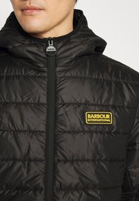 Černá vycpaná bunda s kapucí, s horizontálním prošíváním, předním zipem a žlutým logem "Barbour International".