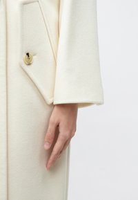 Kermansävyinen villatakki, jossa on tekstuuri ja suuri kulmikas tasku sekä kultainen nappi. Hiha on leveä ja suora.