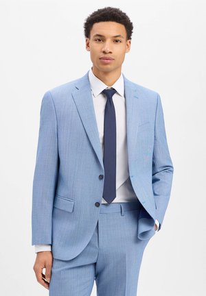 Mann trägt hellblauen Anzug, weißes Hemd und dunkelblaue Krawatte, steht mit einer Hand in der Tasche vor weißem Hintergrund.