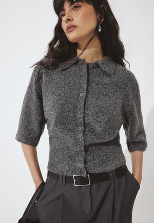 Žena nosi sivý krátky vlnený kardigán s gombíkmi a golierom, tmavé nohavice s čiernym opaskom a drží čiernu tašku.