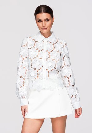 Kvinde iført hvid blomstret blondebluse med knapper og hvid mini-nederdel, stående mod en ensfarvet lys baggrund.