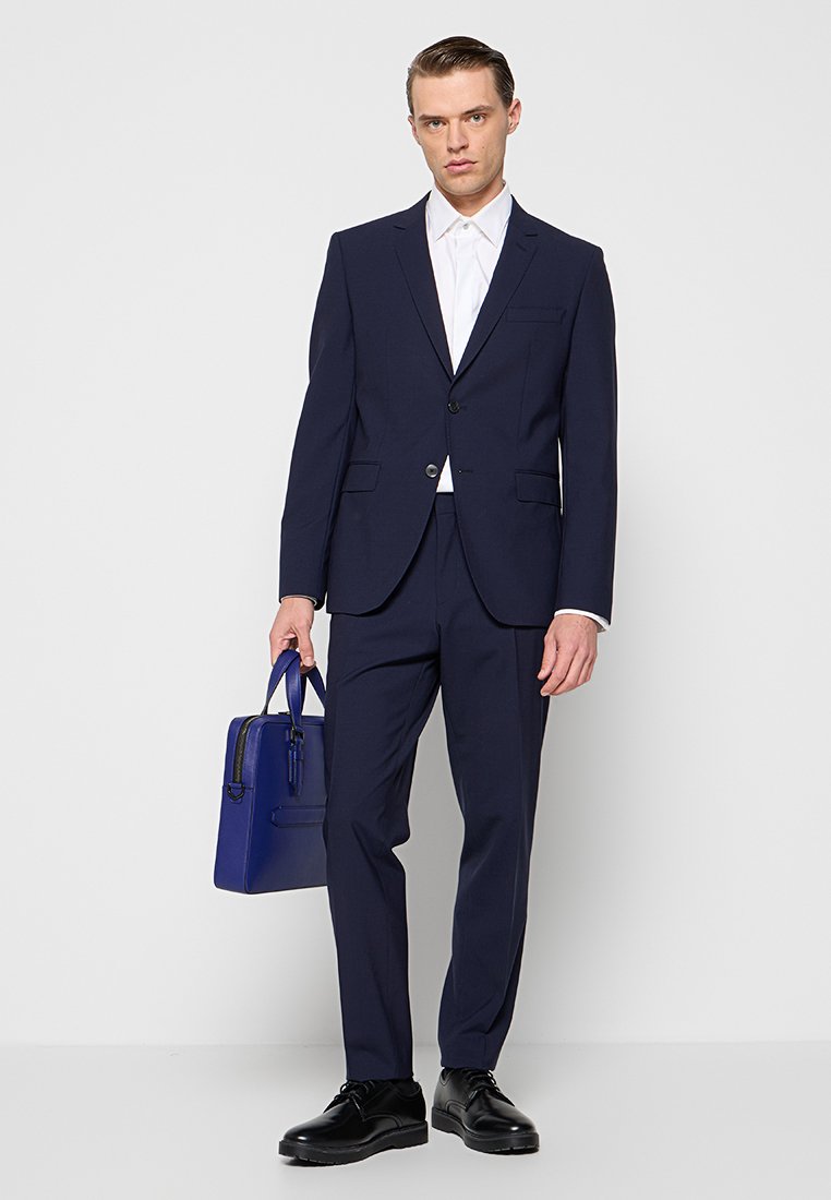 Mann in einem dunkelblauen Anzug, weißem Hemd, schwarzen Schuhen, der eine blaue Lederaktentasche hält und vor einem hellen Hintergrund steht.