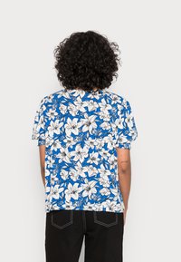 Camisa azul de manga curta com um padrão floral branco. Feita de tecido leve. Gola redonda e um corte descontraído. Barra arredondada.