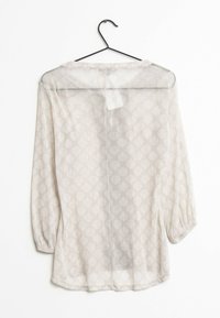 Blouse beige transparente à manches longues avec un motif subtil de feuilles, suspendue sur un cintre noir contre un fond blanc.
