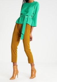 Blusa verde a maniche corte con vita annodata, abbinata a pantaloni giallo senape a gamba stretta. Il modello indossa stivaletti beige a punta.