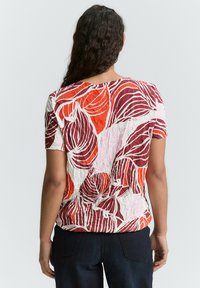 Chemise à manches courtes avec un motif de feuilles rouges et orange sur fond blanc, réalisée en tissu texturé. Vue arrière affichée.