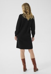 Abito in maglia nera con maniche lunghe, taglio dritto e lunghezza modesta, abbinato a stivali di pelle marrone al ginocchio. Texture liscia.