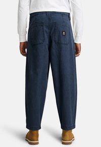 Pantaloni navy blue in stile rilassato, realizzati in tessuto resistente, presentano due tasche posteriori e una piccola etichetta con logo. Indossati con stivali marrone chiaro.