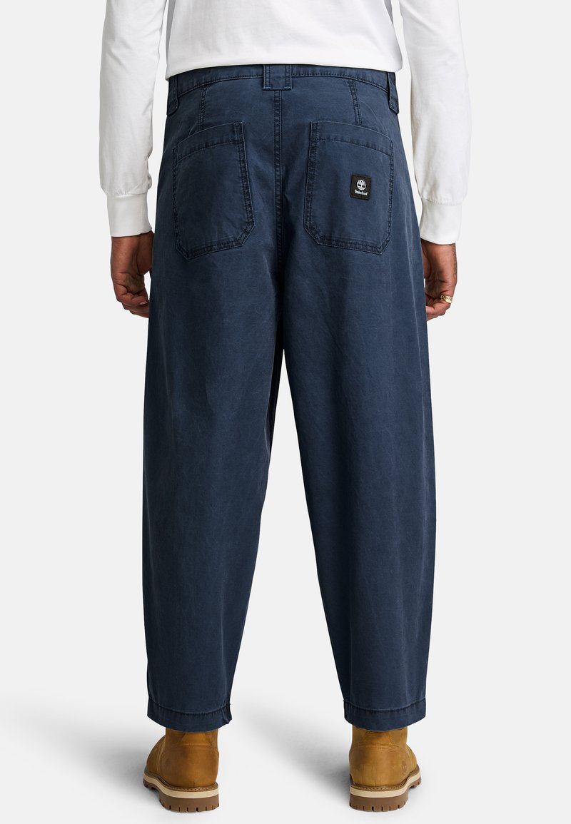 Marineblaue, lässig geschnittene Hose aus robustem Material, ausgestattet mit zwei Gesäßtaschen und einem kleinen Logo-Patch. Getragen mit hellbraunen Stiefeln.