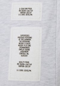 Deux étiquettes en tissu blanches cousues sur un tissu gris, le texte inclut G-STAR RAW PROD ainsi que des détails sur l’entretien et le développement du vêtement.