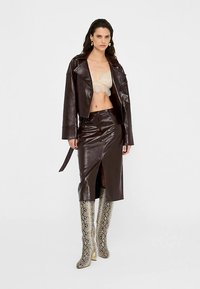 Veste courte en cuir synthétique marron et jupe assortie à fente haute, accompagnées d'un haut en dentelle crème. Bottes montantes à motif serpent complètent la tenue.