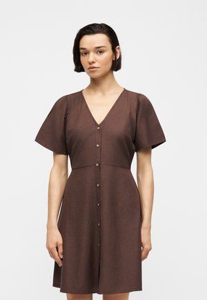Žena s krátkymi tmavými vlasmi, oblečená v hnedých krátkych rukávoch s gombíkmi a V-výstrihom, stojaca pred jednoduchým svetlým pozadím.