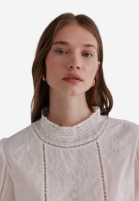 Blusa bianca ricamata con colletto in pizzo, caratterizzata da motivi floreali e tagli verticali lungo la parte anteriore e le maniche. Tessuto morbido.