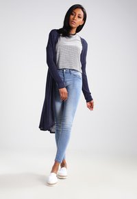 Larga chaqueta de punto azul marino sobre una blusa de rayas gris y blanca, combinada con jeans ajustados azul claro y zapatos de esparto blancos. Texturas suaves en toda la vestimenta.