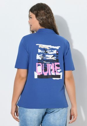 Kobieta w niebieskiej koszulce polo z krótkim rękawem z grafiką latarni morskiej, mew i słowem "DUNE" w różowym kolorze na plecach.