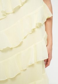 Svetlo rumena obleka z večplastnimi naborki in mehko, prozorno teksturo. Tkanina deluje lahka, z nežnimi, tekočimi robovi.