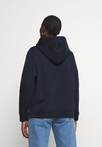 Felpa blu navy con cappuccio regolabile, polsini a coste e tessuto testurizzato. Indossata con jeans azzurri chiari, che mostrano una vestibilità rilassata.