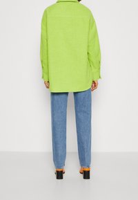 Grüne Fleece-Hemdjacke mit lockerer Passform, langen Ärmeln und Knopfdetails, kombiniert mit hellblauen Jeans und schwarzen High Heels.